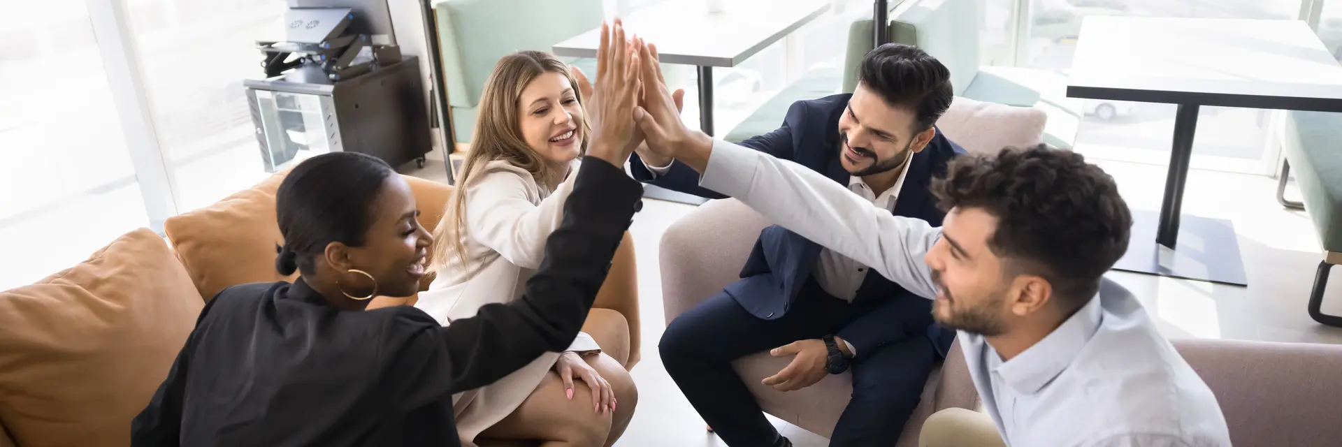 Eine Gruppe von vier Personen in einem modernen Büro feiert gemeinsam einen Erfolg. Sie geben sich gegenseitig High-Fives und wirken glücklich und motiviert. Der Fokus liegt auf der positiven Teamdynamik und Zusammenarbeit. Ein Laptop und Notizen sind auf dem Tisch sichtbar.