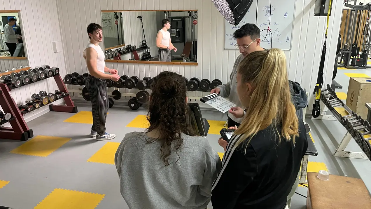 Eine Gruppe von Personen in einem Fitnessstudio. Zwei Frauen und ein Mann stehen zusammen und schauen auf Notizen. Im Hintergrund hebt ein weiterer Mann Gewichte, während ein Lichtsetup auf ihn ausgerichtet ist. Die Umgebung ist mit Hanteln und Fitnessgeräten ausgestattet.