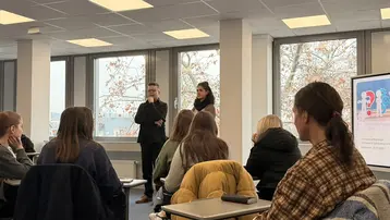 Eine Gruppe von Menschen sitzt in einem Klassenzimmer und hört einem Vortrag zu. Zwei Personen stehen vorne und präsentieren. Im Hintergrund sind Fenster zu sehen, durch die Bäume und eine graue Himmelsszene zu erkennen sind.