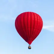 Ein leuchtend roter Heißluftballon schwebt hoch am Himmel. Der Ballon hat eine runde Form und hängt an einem kleinen Korb. Der Hintergrund zeigt einen klaren, blauen Himmel mit wenigen Wolken.