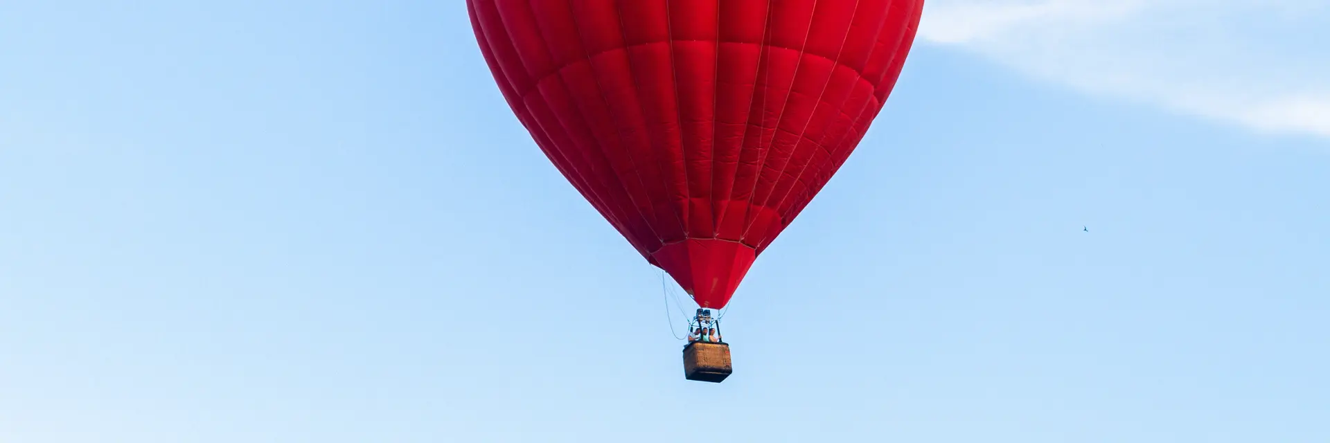 Ein leuchtend roter Heißluftballon schwebt hoch am Himmel. Der Ballon hat eine runde Form und hängt an einem kleinen Korb. Der Hintergrund zeigt einen klaren, blauen Himmel mit wenigen Wolken.