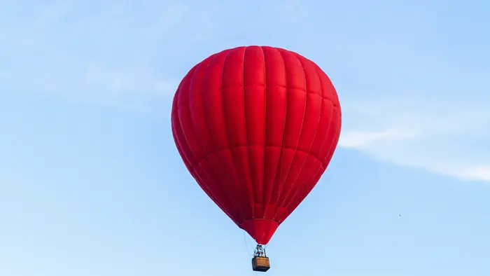 Ein leuchtend roter Heißluftballon schwebt hoch am Himmel. Der Ballon hat eine runde Form und hängt an einem kleinen Korb. Der Hintergrund zeigt einen klaren, blauen Himmel mit wenigen Wolken.