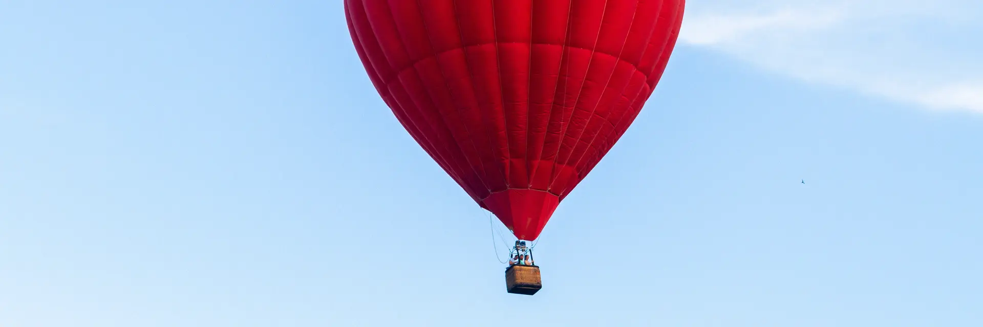 Ein leuchtend roter Heißluftballon schwebt hoch am Himmel. Der Ballon hat eine runde Form und hängt an einem kleinen Korb. Der Hintergrund zeigt einen klaren, blauen Himmel mit wenigen Wolken.