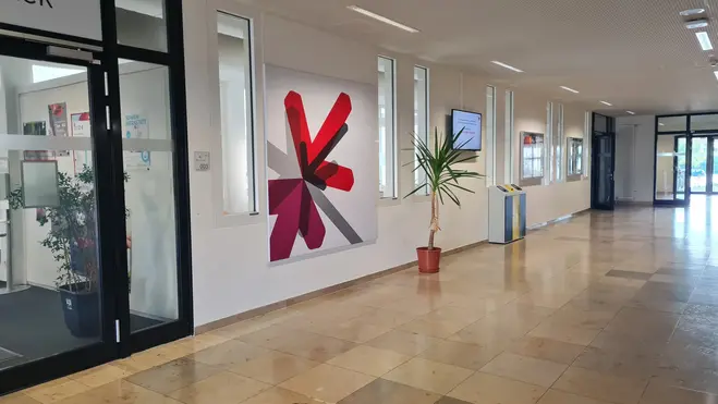 Eine helle Fluransicht mit einem Empfangsbereich auf der linken Seite. An der Wand hängt ein großes rotes und graues Kunstwerk in Form eines stilisierten Buchstabens. Rechts steht ein Topf mit einer grünen Pflanze. Neben dem Empfang sind Informationsschilder sichtbar.