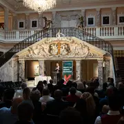 Ein Redner steht auf einer erhobenen Bühne in einem prächtigen Saal mit einer Treppe. Die Zuschauer sitzen im Vordergrund. Im Hintergrund ist ein Plakat mit dem Text „Hat’s im Golden Business?“ zu sehen. Der Raum ist elegant mit kunstvollen Details dekoriert.