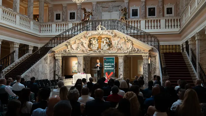 Ein Redner steht auf einer erhobenen Bühne in einem prächtigen Saal mit einer Treppe. Die Zuschauer sitzen im Vordergrund. Im Hintergrund ist ein Plakat mit dem Text „Hat’s im Golden Business?“ zu sehen. Der Raum ist elegant mit kunstvollen Details dekoriert.