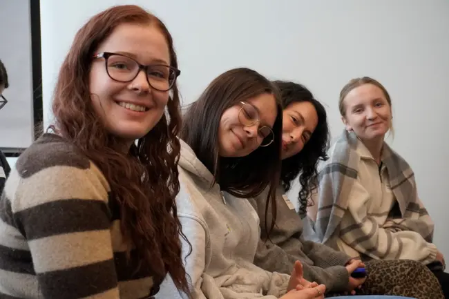 Vier junge Frauen sitzen in einer Reihe und lächeln in die Kamera. Sie tragen lässige Kleidung und wirken freundlich und entspannt. Der Hintergrund ist neutral gehalten.