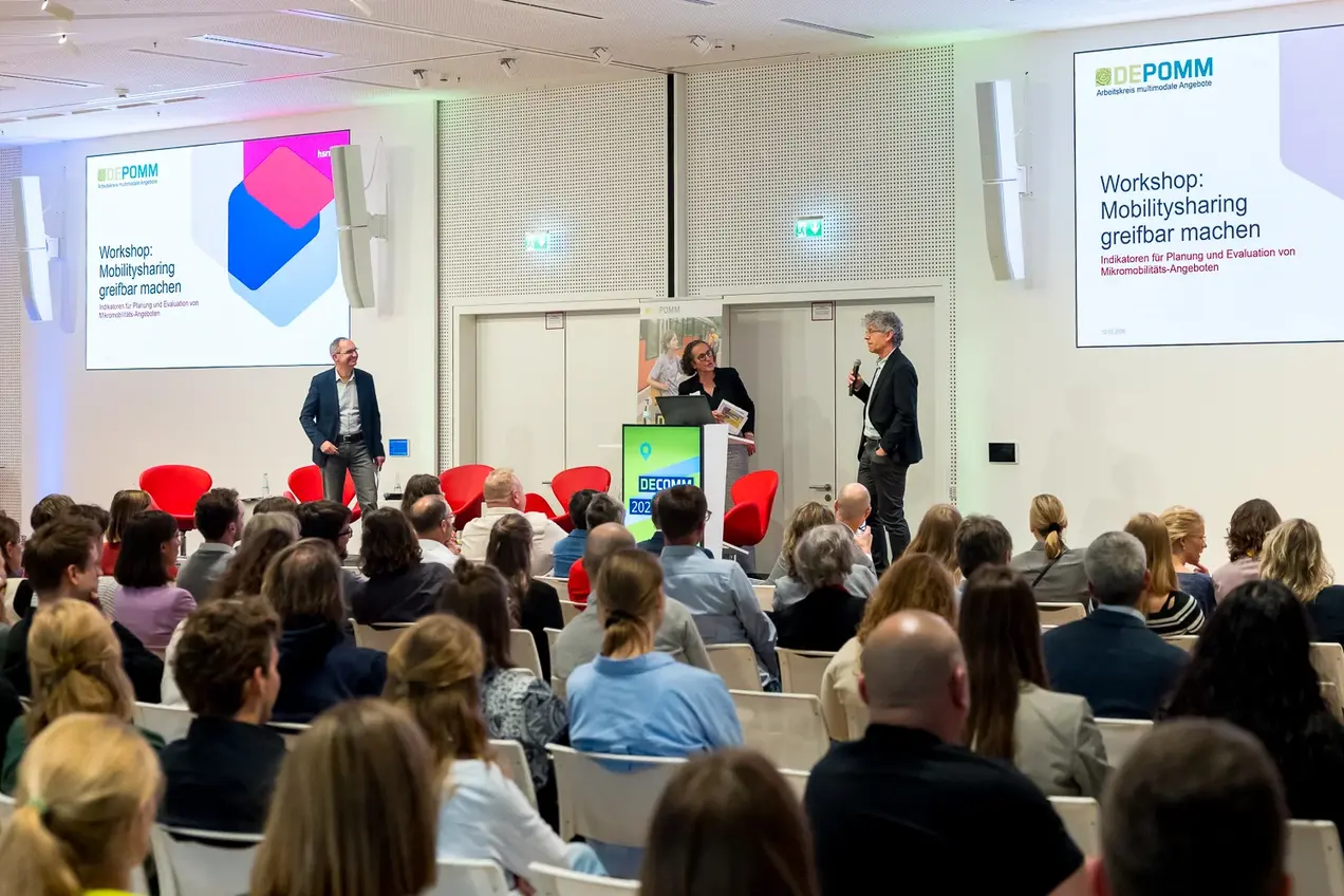 Ein Workshop zu Mobilitätsangeboten findet statt. Auf der Bühne stehen zwei Referenten, während ein Publikum auf Stühlen sitzt und aufmerksam zuhört. Eine Präsentation zum Thema "Mobilitysharing greifbar machen" wird angezeigt.