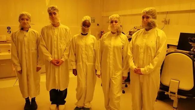 Fünf Personen in Schutzkleidung, einschließlich Hauben und Mundschutz, stehen nebeneinander in einem Labor mit gelblichem Licht. Alle tragen weiße Kittel und spezielle Schuhe, was auf eine sterile Umgebung hinweist.