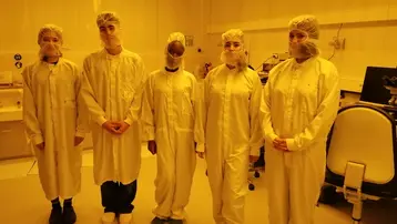 Fünf Personen in Schutzkleidung, einschließlich Hauben und Mundschutz, stehen nebeneinander in einem Labor mit gelblichem Licht. Alle tragen weiße Kittel und spezielle Schuhe, was auf eine sterile Umgebung hinweist.