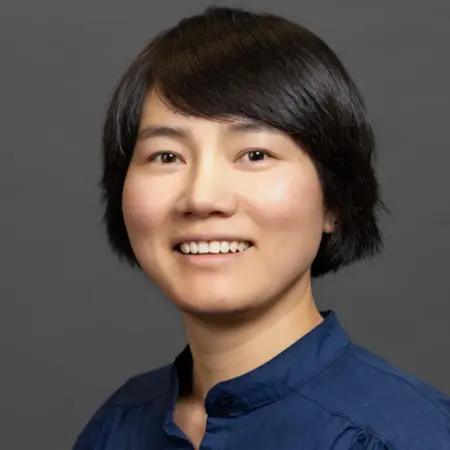 Ein Porträt einer jungen Frau mit schulterlangen, schwarzen Haaren. Sie trägt ein blaues Hemd und lächelt freundlich. Der Hintergrund ist grau, was den Fokus auf ihr Gesicht lenkt.