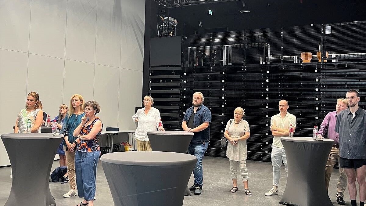 Die Teilnehmenden stehen an Stehtischen und betrachten die Ausstellungsstücke in einer Videopräsentation 