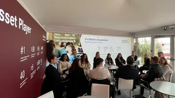 Eine Gruppe von Menschen sitzt an einem Tisch in einem Besprechungsraum. Im Hintergrund ist eine Präsentation zu sehen, die die "Partneria Foundation" und deren Ziel, bessere Ergebnisse zu erzielen, hervorhebt. Der Fokus liegt auf der Interaktion und dem Austausch innerhalb der Gruppe.