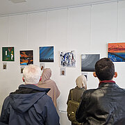 Besucher:innen betrachten die Bilder der Ausstellung, die vor einer weißen Wand hängen.