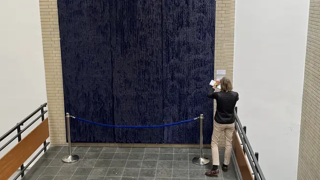 Eine Person steht vor einem großen, blauen Vorhang, der eine Wand verdeckt. Neben der Person sind Absperrband und ein Informationsschild sichtbar. Die Szene befindet sich in einem Innenraum mit Fliesenboden.
