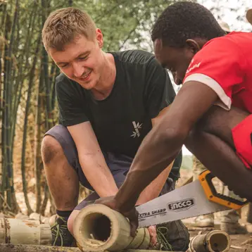 Two men are working together outdoors, cutting bamboo canes. The man on the left is smiling, while the other is concentrating on his work with a hand saw.