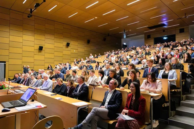 Ein voll besetzter Hörsaal mit zahlreichen Zuhörern, die aufmerksam einer Präsentation folgen. Einige Personen sitzen am Tisch vorne, während im Hintergrund die Zuschauer konzentriert auf den Bildschirm schauen. Die Atmosphäre ist engagiert und interessiert.