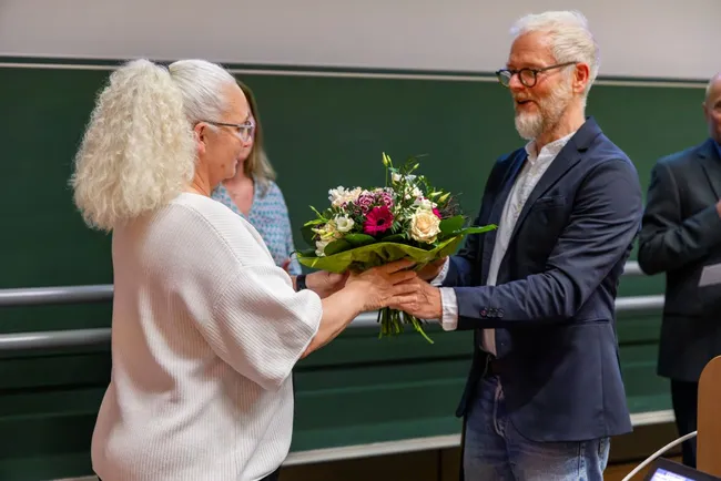 Eine Frau mit lockigem, grauem Haar empfängt einen Blumenstrauß von einem Mann mit Brille und grauen Haaren. Beide lächeln, während sie den Strauß überreichen. Der Hintergrund zeigt eine Gruppe von Personen, die interessiert zuschauen.