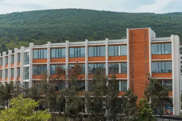 Eine moderne, mehrstöckige Schule mit großen Fenstern und einem orangefarbenen Ziegelfassade. Vor dem Gebäude stehen Bäume, und im Hintergrund ist ein bewaldeter Hügel zu sehen. Der Himmel ist teilweise bewölkt.