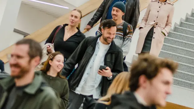 Eine Gruppe von Menschen geht eine Treppe hinunter. Im Vordergrund stehen einige Personen in lässiger Kleidung, während im Hintergrund weitere Personen in formellerer Kleidung zu sehen sind. Die Treppe ist hell erleuchtet und modern gestaltet.