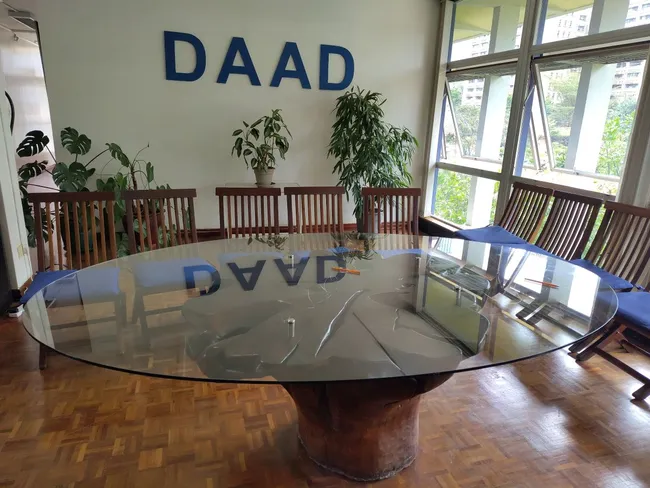 Ein runder Tisch mit einer Glasplatte steht in einem Raum mit Pflanzen und Stühlen. An der Wand ist das Wort „DAAD“ in großen Buchstaben zu sehen. Der Raum hat große Fenster, die viel Tageslicht hereinlassen.