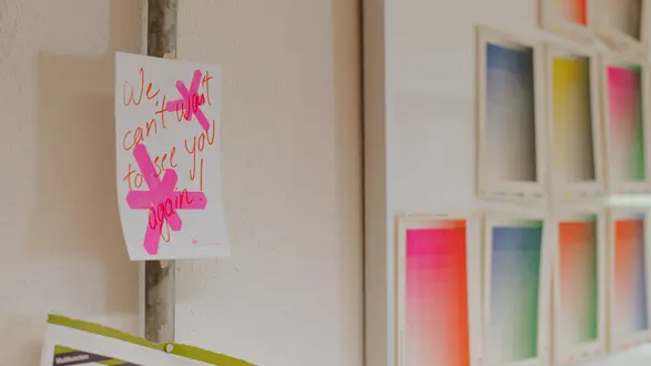 Ein handgeschriebener Zettel hängt an einer Wand und besagt: "We can't wait to see you again!" Teilweise durchgestrichen, um den ersten Satz zu korrigieren. Im Hintergrund sind Farbpaletten mit verschiedenen Farbtönen sichtbar.