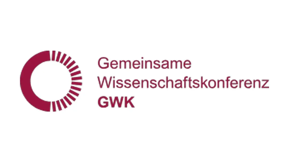 Logo der Gemeinsamen Wissenschaftskonferenz (GWK) in rot und weiß. Die Darstellung zeigt einen stilisierten Kreis mit einem inneren Loch und radialen Linien, begleitet von dem Text „Gemeinsame Wissenschaftskonferenz GWK“.