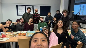 Eine Gruppe von 15 Personen sitzt an einem Tisch, umgeben von Tellern mit Essen und Getränken. Sie lächeln und posieren für ein Foto, während einige die Kamera direkt anschauen. Der Raum wirkt freundlich und gesellig. Die Stimmung ist entspannt und fröhlich.