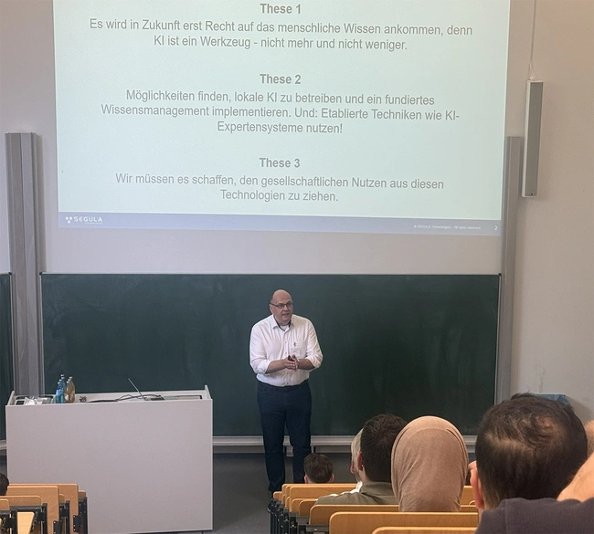 Ein Referent präsentiert in einem Seminarraum mit einer Tafel im Hintergrund. Auf der Tafel sind drei Thesen zur Rolle der KI im Wissensmanagement abgebildet. Der Raum ist mit Stühlen und Tischen ausgestattet, Anwesende hören aufmerksam zu.