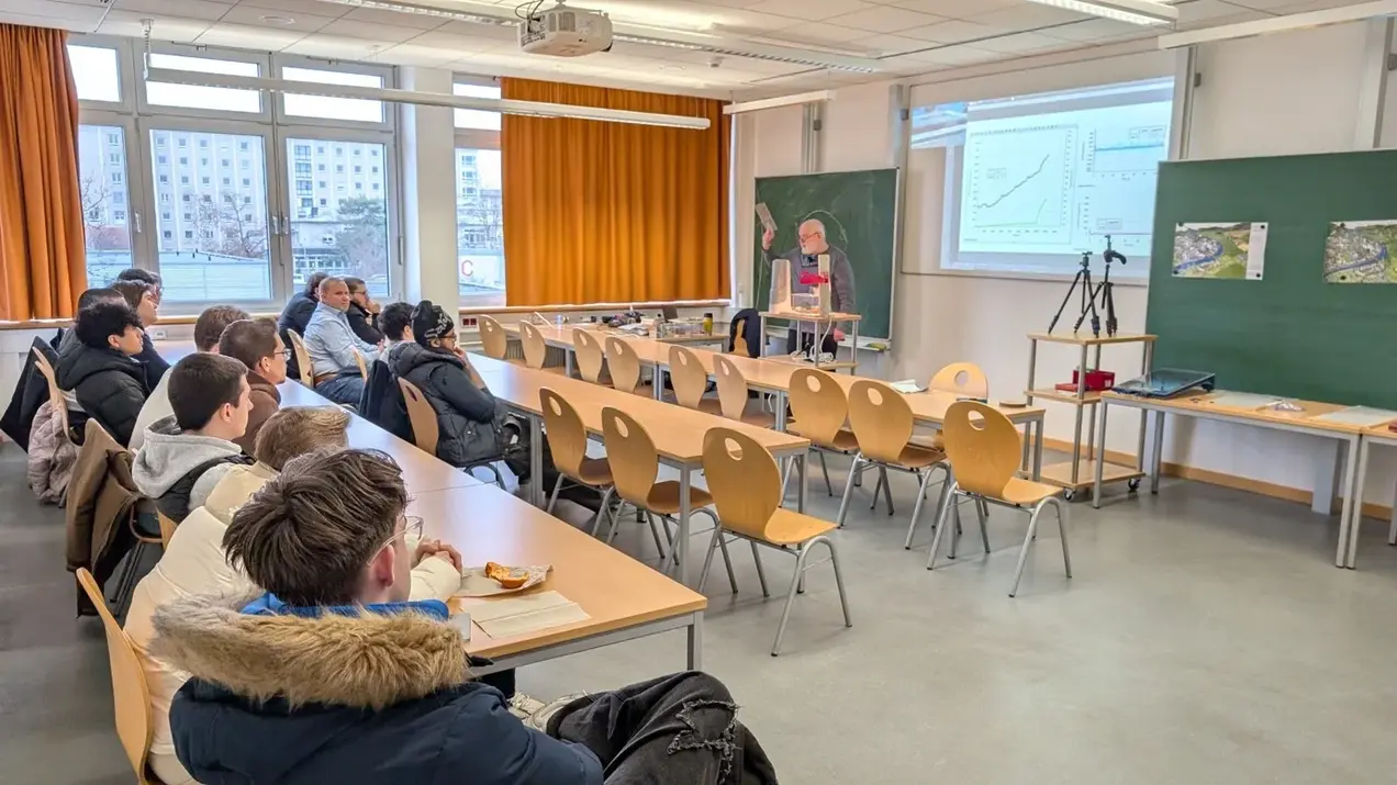 Ein Lehrer präsentiert Daten an einer Projektionsfläche in einem Klassenraum. Die Klasse besteht aus mehreren Schülern, die aufmerksam zuhören. Im Hintergrund sind Tafeln und Fenster zu sehen, die für Tageslicht sorgen.