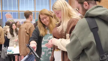 Drei Personen stehen um einen Tisch und betrachten gemeinsam einen Bildschirm. Eine Frau zeigt auf etwas auf dem Bildschirm, während die andere Frau und ein Mann aufmerksam schauen. Im Hintergrund sind weitere Menschen und Tische zu sehen.