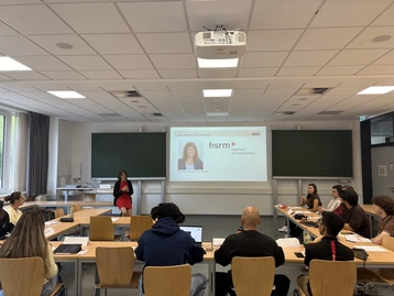 Eine Frau steht vorne in einem Klassenraum und präsentiert etwas auf einer Leinwand. Der Raum ist mit Tischen und Stühlen ausgestattet, und einige Studierende sind auf das Geschehen fokussiert. An der Wand sind zwei digitale Tafeln sichtbar.