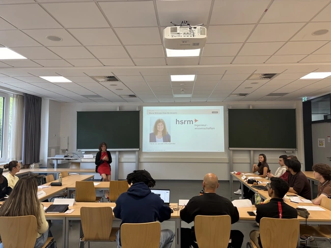 Eine Frau steht vorne in einem Klassenraum und präsentiert etwas auf einer Leinwand. Der Raum ist mit Tischen und Stühlen ausgestattet, und einige Studierende sind auf das Geschehen fokussiert. An der Wand sind zwei digitale Tafeln sichtbar.