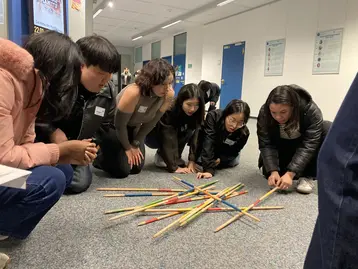 Eine Gruppe von sechs Personen beugt sich über einen bunten Haufen dünner Stäbe auf dem Boden. Sie scheinen konzentriert zu sein und arbeiten gemeinsam an einer Aktivität oder einem Spiel, das Geschicklichkeit erfordert. Der Raum ist hell und modern.