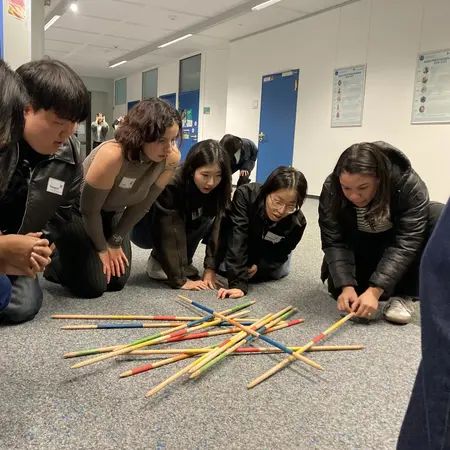 Eine Gruppe von sechs Personen beugt sich über einen bunten Haufen dünner Stäbe auf dem Boden. Sie scheinen konzentriert zu sein und arbeiten gemeinsam an einer Aktivität oder einem Spiel, das Geschicklichkeit erfordert. Der Raum ist hell und modern.