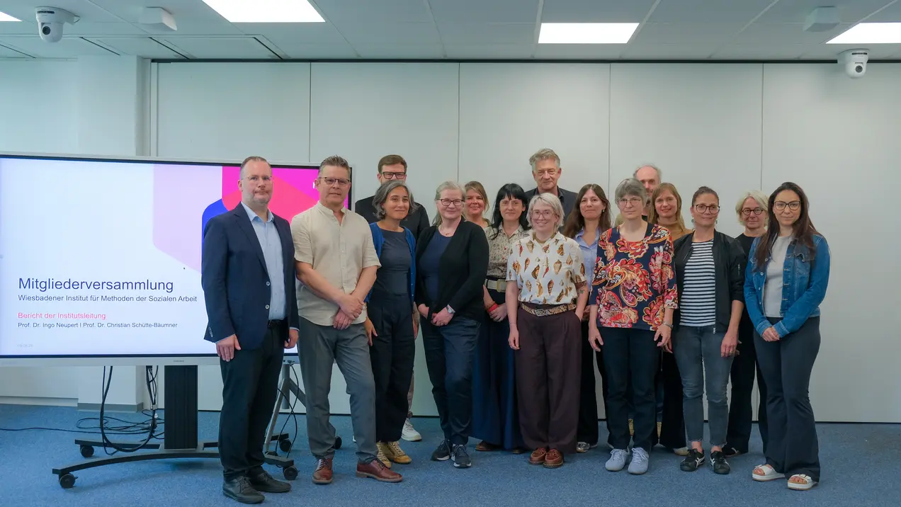 Eine Gruppe von 17 Personen steht zusammen in einem modernen Raum. Im Hintergrund sieht man einen Bildschirm mit der Aufschrift "Mitgliederversammlung". Die Anwesenden sind vielfältig gekleidet und posieren freundlich für das Foto.
