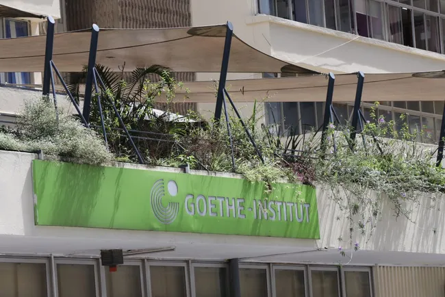 Das Bild zeigt das Goethe-Institut. Es ist ein modernes Gebäude mit einer markanten grünen Fassade, auf der der Schriftzug "GOETHE-INSTITUT" zu sehen ist. Über dem Gebäude wachsen einige Pflanzen auf einem Balkon.