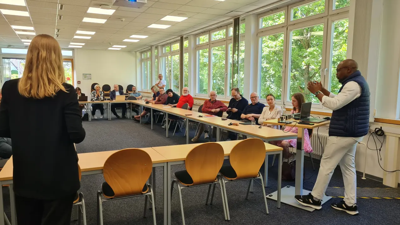 In einem Konferenzraum stehen zwei Personen im Vordergrund. Eine Person spricht, während eine Gruppe von Zuhörern an einem langen Tisch sitzt und aufmerksam zuhört. Große Fenster lassen viel Tageslicht herein. Die Szenen vermitteln eine Atmosphäre des Austauschs und des Lernens.