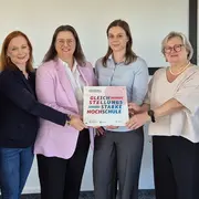 Sechs Frauen stehen in einer Reihe, halten gemeinsam ein Schild mit der Aufschrift „Gleichstellungsstarke Hochschule“. Sie wirken engagiert und festlich, was auf ein wichtiges Ereignis zur Gleichstellung hinweist. Im Hintergrund sind Wände und Fenster zu sehen, die den Raum umgeben.