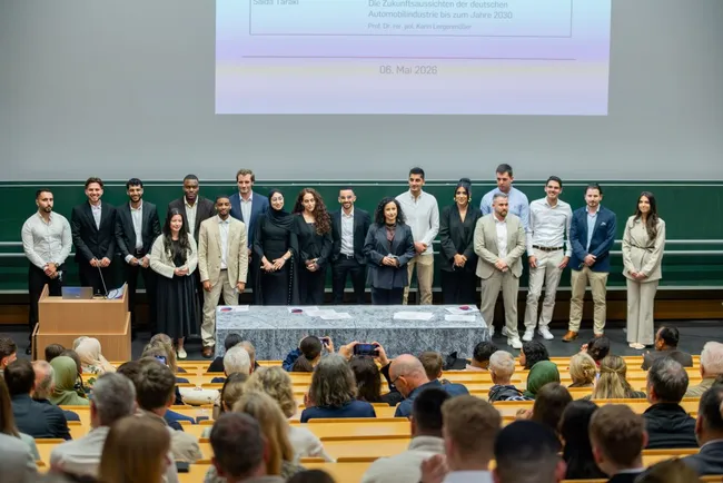 Eine Gruppe von Menschen steht auf der Bühne vor einem Publikum. Sie tragen verschiedene formelle Outfits und posieren für ein Foto. Im Hintergrund ist eine Präsentation auf einer Leinwand zu sehen. Die Atmosphäre wirkt feierlich und offiziell.