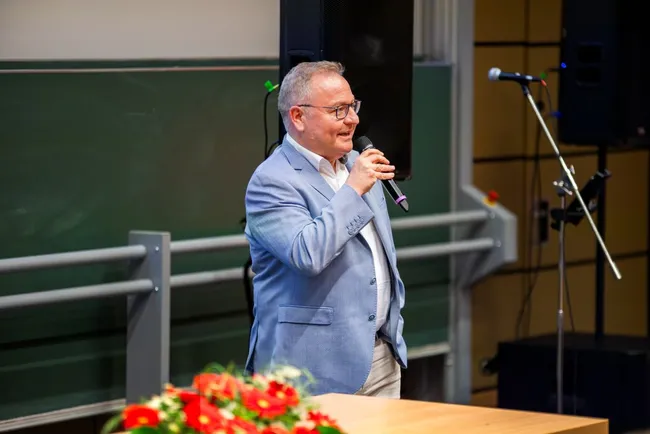 Ein Mann in einem hellen Anzug steht am Rednerpult und spricht in ein Mikrofon. Im Vordergrund sind Blumenarrangements sichtbar. Im Hintergrund befinden sich ein Präsentationsbildschirm und ein Mikrofonstativ.