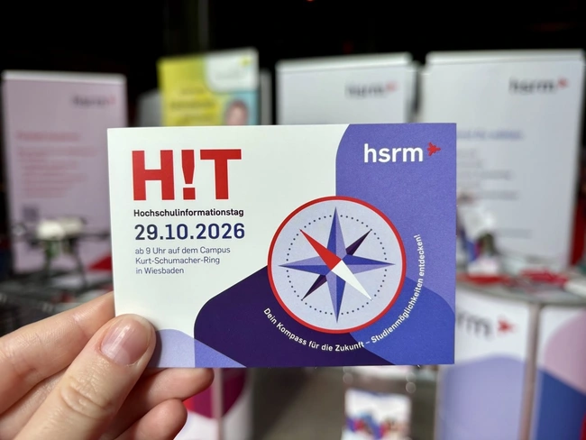 Eine Hand hält eine Informationskarte zum Hochschulinformationstag (HIT) der hsrm. Das Datum ist der 29.10.2026, Beginn um 9 Uhr am Campus Kurt-Schumacher-Ring in Wiesbaden. Das Design zeigt einen Kompass und den Slogan „Der Kompass für die Zukunft – Studienmöglichkeiten entdecken“.