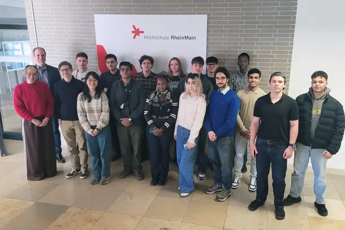 Eine Gruppe von 18 Personen steht vor einem Wandbanner der Hochschule RheinMain. Die Teilnehmenden sind vielfältig gekleidet und lächeln zur Kamera. Die Atmosphäre wirkt freundlich und einladend.