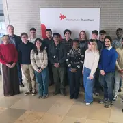 Eine Gruppe von 18 Personen steht vor einem Wandbanner der Hochschule RheinMain. Die Teilnehmenden sind vielfältig gekleidet und lächeln zur Kamera. Die Atmosphäre wirkt freundlich und einladend.