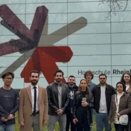 Eine Gruppe von elf Personen steht vor dem Gebäude der Hochschule RheinMain. Im Hintergrund ist das Logo der Hochschule, ein stilisiertes rotes und graues Symbol, deutlich sichtbar. Die Personen sind in verschiedenen Kleidungsstilen gekleidet und lächeln in die Kamera.