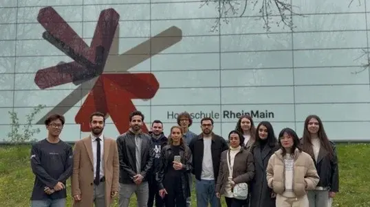 Eine Gruppe von elf Personen steht vor dem Gebäude der Hochschule RheinMain. Im Hintergrund ist das Logo der Hochschule, ein stilisiertes rotes und graues Symbol, deutlich sichtbar. Die Personen sind in verschiedenen Kleidungsstilen gekleidet und lächeln in die Kamera.