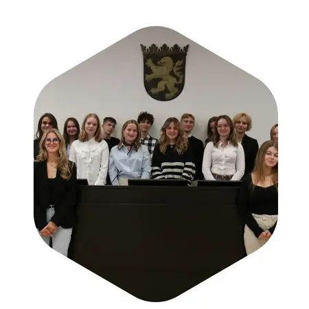 Eine Gruppe von jungen Erwachsenen steht vor einer Wand mit einem Wappen. Sie sind in formeller Kleidung gekleidet und lächeln in die Kamera. Die Szene vermittelt Gemeinschaft und Anlass, möglicherweise eine Veranstaltung oder ein Treffen.