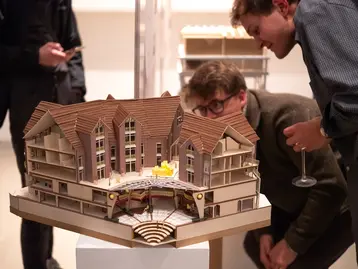 Two people are looking at a detailed architectural model of a building with several floors and a large entrance area. One of the model elements is highlighted in color. In the background, other people can be seen also looking at the model.