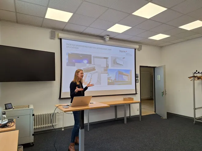 Eine Person präsentiert vor einer Leinwand in einem Seminarraum. Auf der Leinwand sind Bilder und Informationen über das Studium der Ingenieurwissenschaften zu sehen. Der Raum ist hell und modern eingerichtet, mit einem Tisch und einem Laptop im Vordergrund.