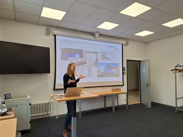 Eine Person präsentiert vor einer Leinwand in einem Seminarraum. Auf der Leinwand sind Bilder und Informationen über das Studium der Ingenieurwissenschaften zu sehen. Der Raum ist hell und modern eingerichtet, mit einem Tisch und einem Laptop im Vordergrund.